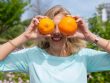 A smiling woman playfully holding oranges in a sunny park setting.